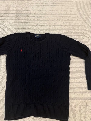Mörkblå kabelstickad tröja Polo Ralph Lauren - Kabelstickad mörkblå tröja från Polo Ralph Lauren med rund halsringning och röd broderad logga på bröstet. Tröjan har långa ärmar och ribbade muddar vid ärmslut och nederkant. Perfekt för lager på lager och klassisk stil. Xl men passar mig i m,