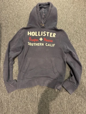 Mörkblå Hollister hoodie med tryck - Mörkblå hoodie från Hollister med stor huva och dragsko. Framtill står det 'HOLLISTER Surfer + Rescue SOUTHERN CALIF' i vitt och rött tryck. Hoodien har en klassisk känguruficka och ribbade muddar. Perfekt för en avslappnad och cool stil.