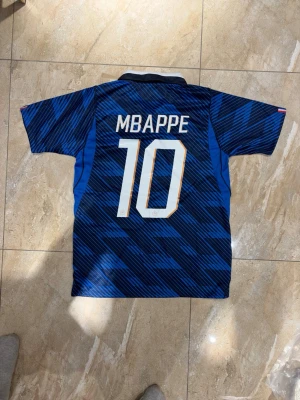 Blå Frankrike fotbollströja Mbappé - Säljer en blå fotbollströja från Nike med Frankrikes landslagsemblem och nummer 10 på bröstet. På ryggen står det Mbappé och siffran 10. Tröjan har korta ärmar, vit krage och guldiga detaljer. Perfekt för dig som älskar fotboll och vill stötta Frankrike.
