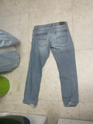 Ljusblå jeans från Nudie Jeans - Säljer ett par ljusblå jeans från Nudie Jeans med klassisk femficksdesign och signatursöm på bakfickorna. Jeansen har raka ben, normal passform och råa, fransiga benslut för en avslappnad vibe. Tillverkade i mjuk bomullsdenim.