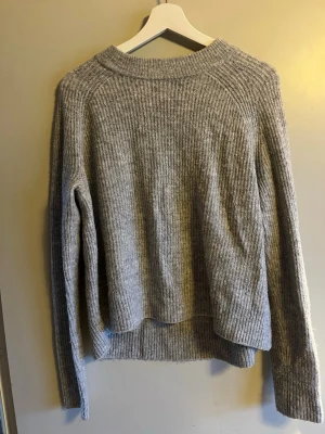 Grå ribbad stickad tröja - Mysig grå stickad tröja med ribbad struktur och rund hals. Tröjan har lång ärm och en något loose passform, perfekt för att hålla värmen under kyliga dagar. Enkel och stilren design som funkar till jeans eller mjukisbyxor.