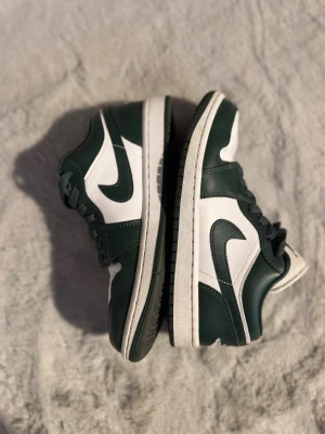 Nike Air Jordan 1 Low gröna/vita - Nike Air Jordan 1 Low sneakers i mörkgrönt och vitt skinn. Klassisk siluett med rund tå, snörning och platt sula. Ikonisk Jumpman-logga på tungan och broderad Air Jordan Wings på hälen. Perfekt för dig som vill ha en clean och sportig look. I mycket bra skick, kan ha små skavanker men märks knappt och påverkar inte användningen 