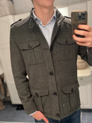 Grå overshirt/peacoat från Peacocks - Snygg grå rock från Peacocks i storlek medium. Jackan har fyra stora fickor med lock och knappar, axelklaffar och klassisk krage. Materialet är ullblandning med en stilren, mörkgrå ton. Perfekt för dig som vill ha en clean och tidlös look.