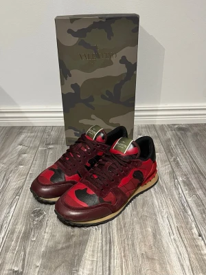 Valentino Rockrunner röda sneakers - Säljer ett par Valentino Rockrunner sneakers i rött och svart med camouflage-mönster. Skorna har detaljer i mocka och skinn, platt sula och snörning. Baksidan har coola nitar och svarta hälpartier. Kommer med originalkartong i camo-print.