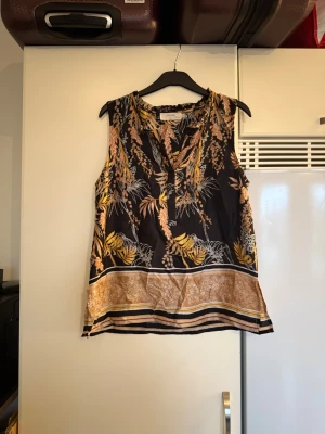 Svart blommig linnetopp från Cream - Svart linnetopp från Cream med blommigt mönster i gult, beige och vitt. Toppen är ärmlös och har en v-ringad hals. Materialet är mjukt och lätt, perfekt för varma dagar. Snygga detaljer med mönstrad kant nedtill som ger extra stilpoäng. Storlek 40. Men är liten i storlek. Mycket fint skick, jag hittar inga synliga anmärkningar eller defekter. 
