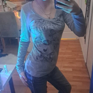 Grå långärmad topp med silvertryck - Säljer en grå långärmad topp med snyggt silverfärgat tryck och blommönster framtill. Toppen har en lätt v-ringning och sitter figurnära. Perfekt för dig som gillar detaljer och vill sticka ut lite extra.