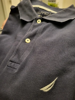 Marinblå pikétröja från Nautica S - Klassisk marinblå pikétröja från Nautica i storlek S. Tröjan har korta ärmar, krage och två vita knappar framtill. Materialet är pikéstickad bomull och på bröstet finns Nauticas ikoniska logga broderad i vitt. Perfekt för en clean och stilren look.