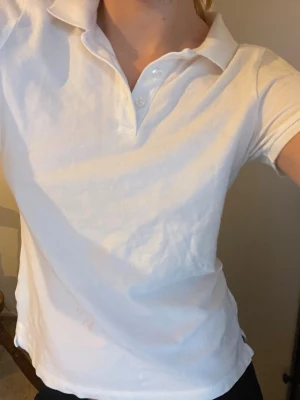 Vit polotopp med krage och knappar - Klassisk vit polotopp med krage och tre knappar framtill. T-shirten har korta ärmar och är gjord i mjukt bomullstyg. Enkel och stilren modell som passar till det mesta.