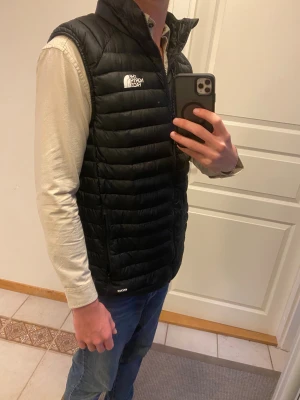 Svart dunväst från The North Face - Säljer en svart dunväst från The North Face med 800 fill power. Västen har quiltad design, hög krage och dragkedja framtill. Perfekt för lager-på-lager och har en clean look med logga på bröstet. Passar dig som gillar street och outdoor.