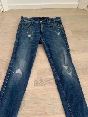 Replay jeans blå  - Snygga blå jeans från Replay med coola slitningar och detaljer på både fram- och bakfickor. Klassisk femficksmodell med knapp och dragkedja. Jeansen har en normal passform och är tillverkade i slitstarkt denimtyg.