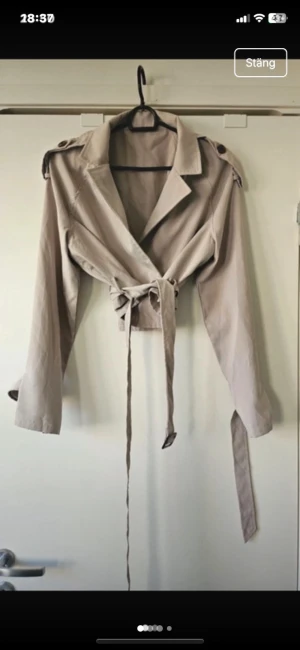 Beige croppad trenchcoat med bälte - Snygg croppad trenchcoat i beige med klassisk krage, axelklaffar och långa ärmar. Jackan har ett omlottband i midjan som knyts och ger en cool siluett. Perfekt att styla med jeans eller kjol för en trendig look.