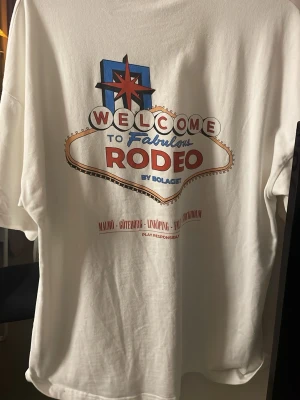 Vit Rodeo t-shirt från Bolaget - Vit t-shirt med tryck 'RODEO' fram och stort Vegas-inspirerat motiv på ryggen. Oversized passform och rund hals. Materialet är mjuk bomull och trycket har röda, blå och beige detaljer. Perfekt för dig som gillar statement-plagg och streetstyle.