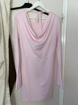 Ljusrosa långärmad klänning med draperad rygg - Supersöt ljusrosa kortklänning med långa ärmar och snygg draperad rygg med knytband. Klänningen har en enkel, stilren framsida och är gjord i ett mjukt, stretchigt material som sitter bekvämt. Perfekt för dig som gillar en feminin och trendig look.