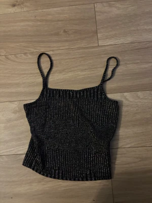 Glittrig svart linnetopp med smala band - Säljer en snygg svart linnetopp med glittriga detaljer och smala axelband. Toppen har ett ribbat mönster. Är använd 1 gång 