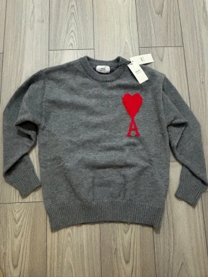 Grå stickad tröja från Ami Paris - Grå stickad tröja från Ami Paris med röd hjärtlogga och bokstaven A på bröstet. Klassisk rund halsringning, ribbade muddar och nederkant. Perfekt för dig som gillar stilrena plagg med en twist.
