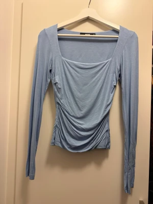 Ljusblå långärmad topp från BikBok - Supersnygg ljusblå långärmad topp från BikBok med fyrkantig ringning och draperad front. Tunn och mjuk i materialet, perfekt för lager-på-lager eller som den är. Sitter figurnära och har en stilren look som passar till jeans eller kjol.