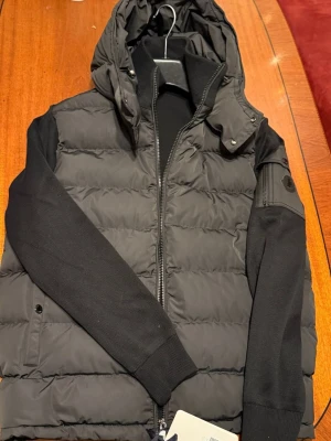 Svart Moncler pufferjacka med huva - Säljer en svart pufferjacka från Moncler med ribbade ärmar och två dragkedjeförsedda fickor. Jackan har en stor huva och Moncler-logga på ärmen. Materialmix med dunfylld kropp och stickade detaljer på ärmar och muddar. Perfekt för dig som vill ha en clean och exklusiv look.