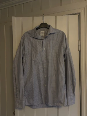 Blårutig skjorta Oscar of Sweden - Slim fit skjorta från Oscar of Sweden med små blå rutor på vit botten. Klassisk krage, långa ärmar och vita knappar framtill. Tillverkad i bomull med tvåfold-kvalitet för extra komfort och stil. Perfekt för dig som gillar en clean och fräsch look.