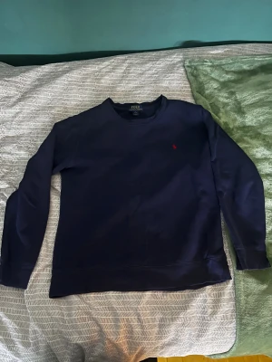 Marinblå tröja från Polo Ralph Lauren - Snygg marinblå långärmad tröja från Polo Ralph Lauren med klassisk crewneck och röd broderad logga på bröstet. Mjuk och skön bomullskvalitet som passar perfekt till jeans eller chinos. Enkel stil med ribbade muddar vid ärmslut och nederkant. Är också liten i storlek 