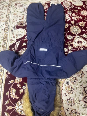 Mörkblå täckbyxa från Reima med fuskpäls storlek 104 barn - Mörkblå täckbyxa från Reima med huva och fuskpälsdetalj. Byxan har resår i midjan och vid benslut, samt reflexdetalj på ryggen. Perfekt för kalla vinterdagar och snölek. Materialet är slitstarkt och vattenavvisande syntet. 