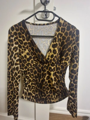 Leopardmönstrad omlottopp med v-ringning - Snygg omlottopp med v-ringning och leopardmönster i bruna och beige toner. Toppen har långa ärmar och lätt rynkade detaljer vid sidorna för en cool siluett. Mjuk och stretchig polyester som sitter skönt och ger en slim passform. Även jättefin skick!