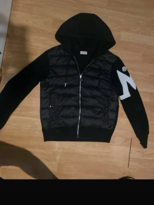 Svart Moncler pufferjacka med huva - Svart pufferjacka från Moncler med huva och hel dragkedja framtill. Jackan har stickade ärmar med vit zigzag-detalj på ena armen och Moncler-logga. Kroppen är quiltad och tillverkad i syntetmaterial, medan ärmarna är stickade. Snygg och sportig look med coola detaljer.