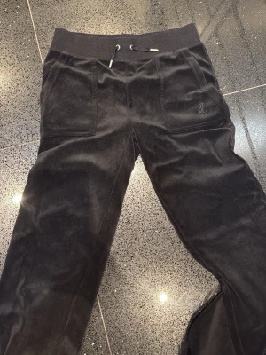 Svarta Juicy Couture byxor - Säljer ett par svarta Juicy Couture mjukisbyxor i mjuk sammet med bred resårmidja och snörning. Byxorna har två stora fickor fram och en diskret broderad logga på ena fickan. Perfekta för chill dagar eller när du vill vara extra bekväm.