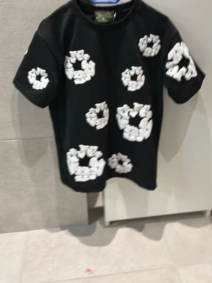 Svart t-shirt med vita broderade cirklar - Cool svart t-shirt från Denim Tears i bomull med unika vita broderade cirkelmönster över hela plagget. Klassisk rund hals och korta ärmar. Perfekt för dig som vill sticka ut med en streetwear-vibe och snygga detaljer.