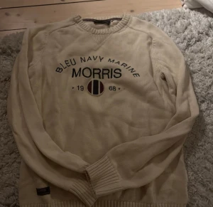 Beige stickad tröja Morris S - Beige stickad tröja från Morris med broderad text och emblem på bröstet. Tröjan har rund hals, ribbade muddar och är långärmad. Perfekt för dig som gillar klassisk stil med en twist. Snygg detalj med randigt band på insidan av kragen. Använd ett väldigt fåtal gånger 