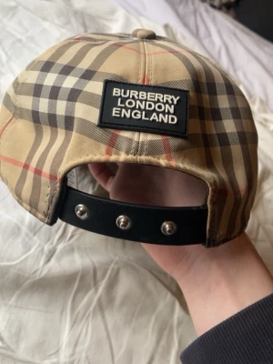 Burberry rutig keps beige L - Säljer en klassisk Burberry keps i beige med ikoniskt rutigt mönster och svart justerbart spänne bak. Märkt med 'Burberry London England' på baksidan. Tillverkad i bomull och polyester, perfekt för dig som vill ha en clean och tidlös accessoar.
