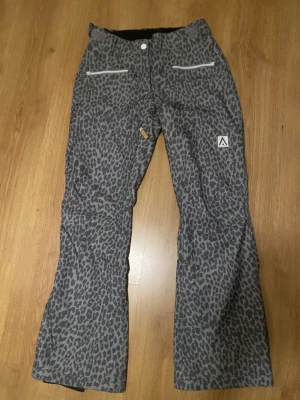 Grå leopardmönstrade täckbyxor - Snygga grå täckbyxor med leopardmönster och två dragkedjefickor framtill. Byxorna har en bootcut-passform och är perfekta för dig som vill sticka ut i backen. Märke med logga på benet och detaljer i vitt och orange.