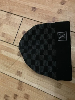 Rutig svart väst från Louis Vuitton - Snygg svart väst från Louis Vuitton med klassiskt rutigt mönster och LV-logga på bröstet. Västen har en stilren design och är tillverkad i ett mjukt stickat material som känns skönt mot kroppen. Perfekt för dig som gillar exklusiva märken och vill sticka ut.