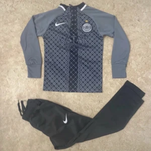 Nike PSG träningströja och byxor - Säljer ett set med Nike PSG träningströja i grått och svart med geometriskt mönster och lång ärm, samt svarta träningsbyxor med vit Nike-logga. Tröjan har PSG-emblem och Qatar Airways-tryck. Perfekt för fotbollsträning eller chill.