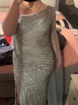 Glittrig silver one shoulder långklänning - Säljer en magisk långklänning i silver med glittriga paljetter och pärlor. Klänningen har ett elegant one shoulder-snitt med transparent släp på ena armen och en snygg sl soppen slits. Perfekt för bal eller nyår! Materialet känns lyxigt och skimrar i ljuset. Använd bara en gång köpte den den kostade 7000kr.
