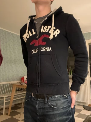 Hollister Zip Up med luva - En sjukt snygg Hollister California zip up med en riktigt najs vintage känsla🤩!  Mörkblå med snyggt broderi. Är i storlek xs(står M på lappen, stämmer ej) och passar någon <170cm. Är 176cm på bilden, så den sitter kort. Var inte rädd att skicka frågor och prisförslag!😁
