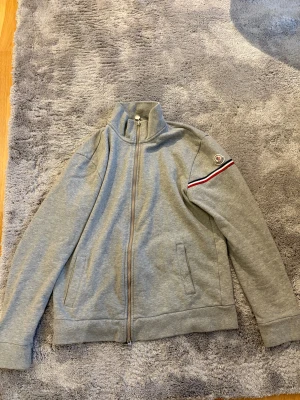 Moncler zip - Snygg grå kofta från Moncler med hel dragkedja och ståkrage. På vänster ärm finns Moncler-logga och en röd, vit och blå rand som detalj. Tröjan har två fickor fram och är tillverkad i mjuk bomullsmix. Perfekt för dig som gillar stilrena och sportiga plagg. Stämplad xl men passar M, köpt på mearchsweden 