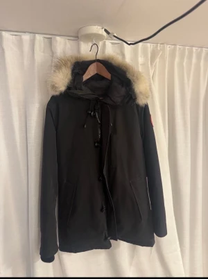 Svart dunjacka Canada Goose med pälskrage - Svart dunjacka från Canada Goose med ikonisk röd logga på ärmen. Jackan har en fluffig pälskant på huvan, dragkedja och knappar framtill samt två stora fickor. Insidan är quiltad och fodrad för extra värme. Perfekt för kalla vinterdagar.