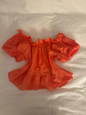 Orange blus - Säljer en orange blus från Samsa Collection med offshoulder och puffiga korta ärmar. Blusen har volangdetaljer vid halsringningen och nederkanten.