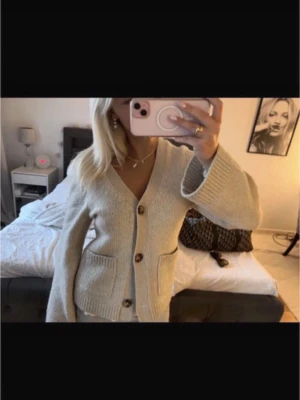 Beige stickad kofta med knappar - Säljer en beige stickad kofta med v-ringad hals och stora bruna knappar framtill. Koftan har två praktiska fickor och långa ärmar med lite loose passform. Perfekt att slänga på sig när det är lite kyligt ute.