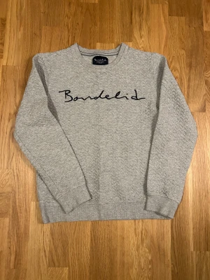 Grå sweatshirt från Bondelid S - Snygg grå sweatshirt från Bondelid i storlek S med svart broderad logga på bröstet. Tröjan har ett diskret vågigt mönster i tyget, rund hals och långa ärmar. Perfekt för dig som gillar stilrena och bekväma plagg med lite extra detalj.
