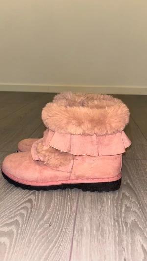 Rosa fluffiga boots med volangdetalj - Supergulliga rosa boots med fluffig pälsimitation runt skaftet och volangdetaljer. Skorna har en grov sula och är gjorda i mockaliknande material, perfekta för att hålla värmen under kyliga dagar. Snyggt och mysigt statement till vintern!