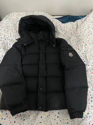 Moncler Vezere Jacka - Säljer en svart pufferjacka från Moncler med stor huva och snygga knappar framtill. Jackan har en ficka med Moncler-logga på ärmen och är riktigt fluffig och varm. Perfekt för kalla dagar och har en cool, modern look. Den är använd i en månad ungefär, inga som helst fel på den. Storlek 1=S