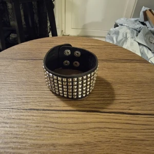 Svart nitarmband med tryckknappar - Säljer ett brett svart armband med flera rader silvriga nitar och två tryckknappar för stängning. Längd 22,5 cm. Armbandet är gjort i skinnimitation i PU och har en cool, edgy vibe som passar perfekt till streetwear eller rockiga outfits.