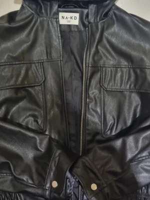 Svart skinnjacka från NA-KD - Snygg svart skinnjacka från NA-KD med dragkedja framtill och två stora fickor med lock. Jackan har en cool, lite loose passform och detaljer med tryckknappar vid ärmsluten. Perfekt för dig som vill ha en edgy stil.