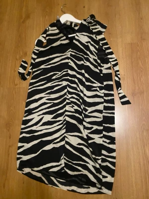 Zebramönstrad långklänning med knyt - Säljer en långklänning med svartvitt zebramönster och v-ringning. Klänningen har knytband vid axeln och ärmlös design, vilket ger en cool och trendig look. 