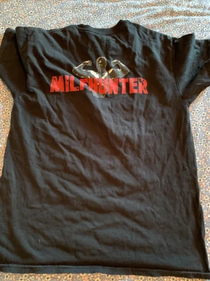 Svart t-shirt med MILFHUNTER-tryck - Svart t-shirt med coolt MILFHUNTER-tryck i rött och en siluett av en flexande person på ryggen. Klassisk rund hals och korta ärmar, perfekt för dig som gillar statement-plagg. Materialet känns som mjuk bomull. Kom med prisförslag om ni tycker priset e för högt!