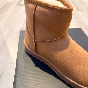 Bruna mockaboots med fårull - Mysiga boots i brun mocka med mjukt, vitt fårullsfoder på insidan. Skorna har en platt sula och rund tå, perfekta för kalla dagar. Klassisk design med synliga sömmar och kort skaft.