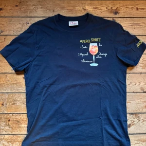 Mörkblå Aperol Spritz t-shirt Saint Barth - Snygg mörkblå t-shirt från MC2 Saint Barth i ekologisk bomull. Framsidan har ett färgglatt Aperol Spritz-tryck med receptdetaljer och glas. Klassisk passform och korta ärmar, perfekt för dig som gillar coola print och sommarkänsla.                    Lite lätt solblekt på ryggen!