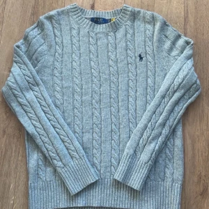 Grå kabelstickad tröja Polo Ralph Lauren - Snygg grå kabelstickad tröja från Polo Ralph Lauren med klassisk rund hals och broderad marinblå logga på bröstet. Tröjan har långa ärmar och breda ribbade muddar vid ärmslut och nederkant. Perfekt för dig som gillar stilren och tidlös design.