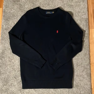Mörkblå stickad tröja från Polo Ralph Lauren - Klassisk mörkblå stickad tröja från Polo Ralph Lauren med rund halsringning och röd broderad logga på bröstet. Tröjan har ribbade muddar vid ärmslut och nederkant, samt långärmad passform. Perfekt för en stilren look.